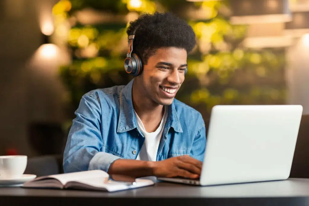 Homem sorrindo e sentado em frente ao computador e um livro aberto em cima da mesa