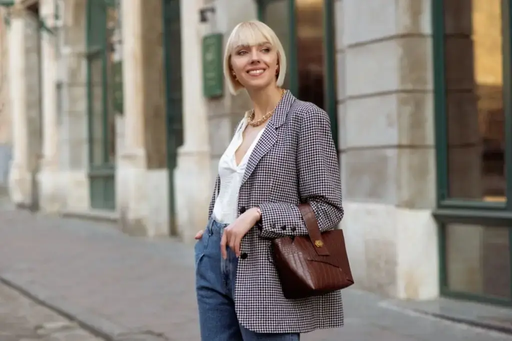 Mulher loira com blazer xadrez, blusa branca e calça jeans sorrindo em rua