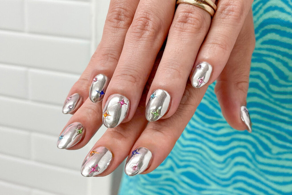 duas mãos com unhas prateadas, com efeito cromado e pedras coloridas