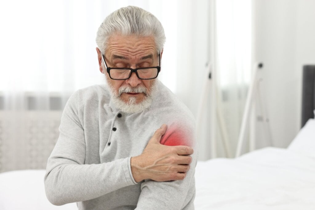Homem idoso com cabelo curto grisalho, usando óculos preto e blusa de frio cinza, sentado na cama com a mão no ombro indicando estar com dor no ombro