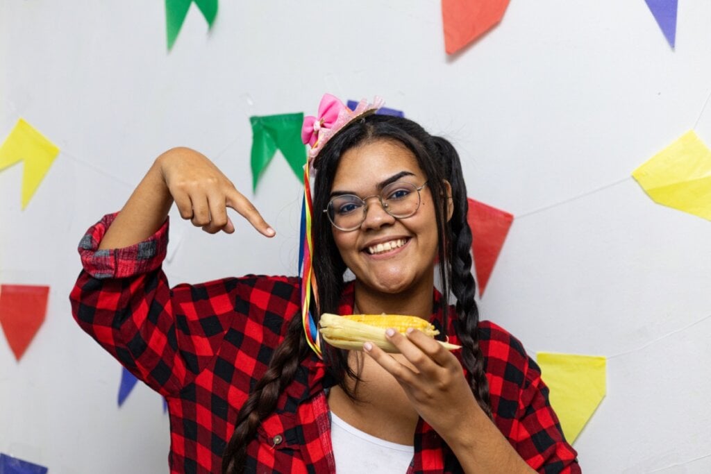 Jovem usando camisa xadrez e adereço de festa junina no cabelo, com o cabelo trançado e usando óculos com um milho na mão e com decoração de festa junina na parede do fundo