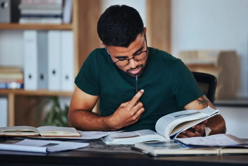 menino de camiseta verde sentado com caneta perto da boca e olhando para livro aberto em cima de mesa 