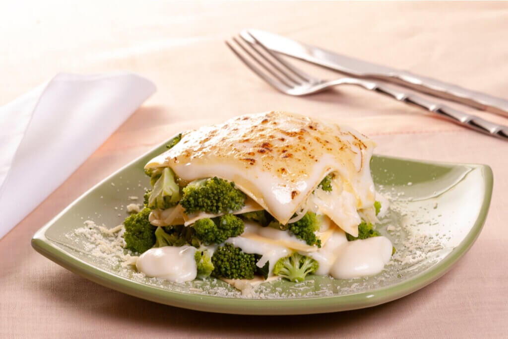 Lasanha de brócolis com molho branco em prato verde, garfo e faca e guardanapo ao fundo