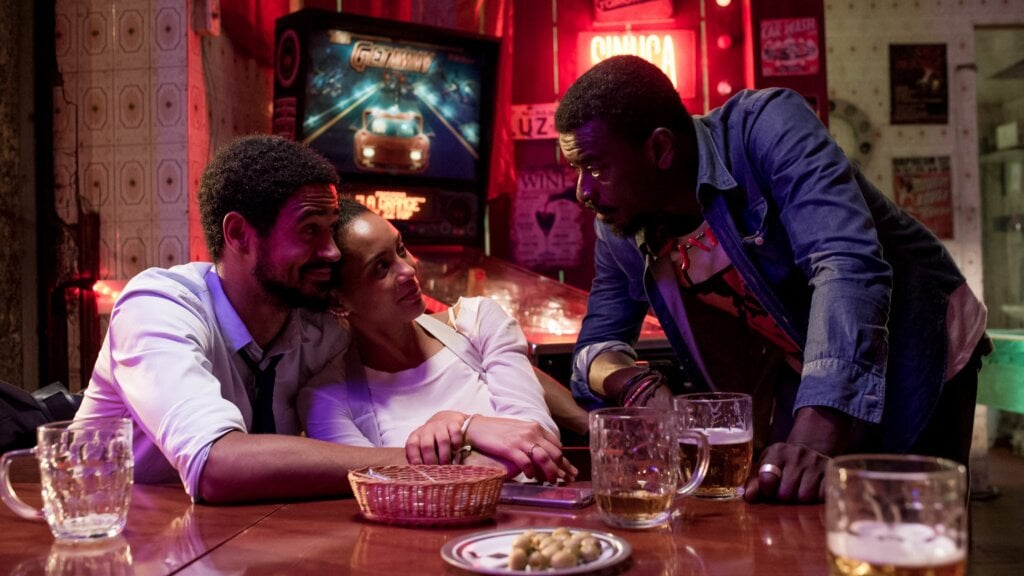 Casal abraçado sentado em mesa de bar, conversando com outro homem que está de pé. Há copos de bebidas e pratos com comida na mesa