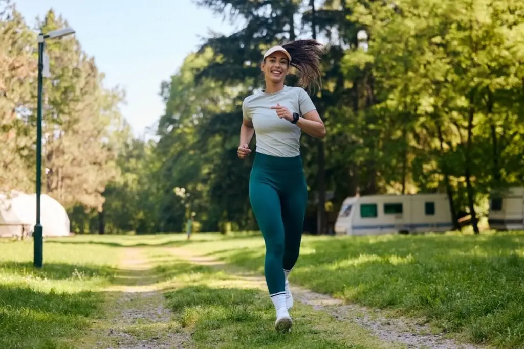 Mulher com o cabelo preso, usando boné, uma camiseta cinza e calça verde e tênis branco correndo ao ar livre