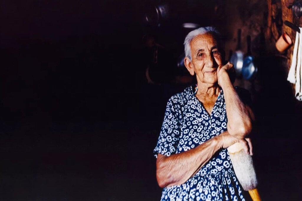 Mulher idosa com um vestido estampado apoiada em um pedaço de madeira apoiando o rosto em uma das mãos