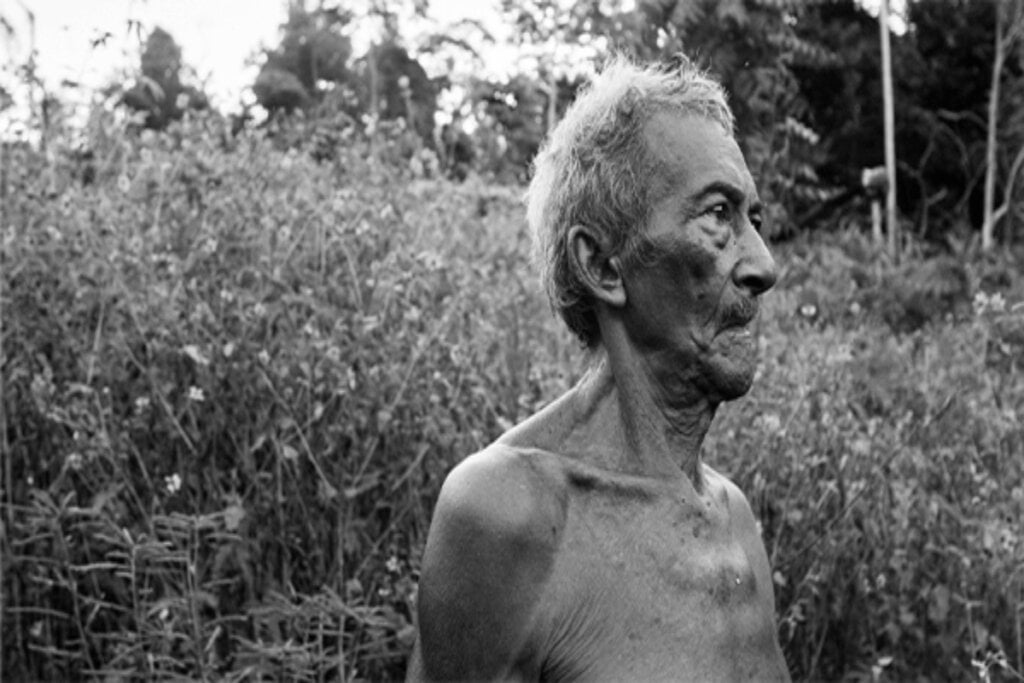 Imagem em preto e branco de um homem isdoso sem camisa no meio da mata