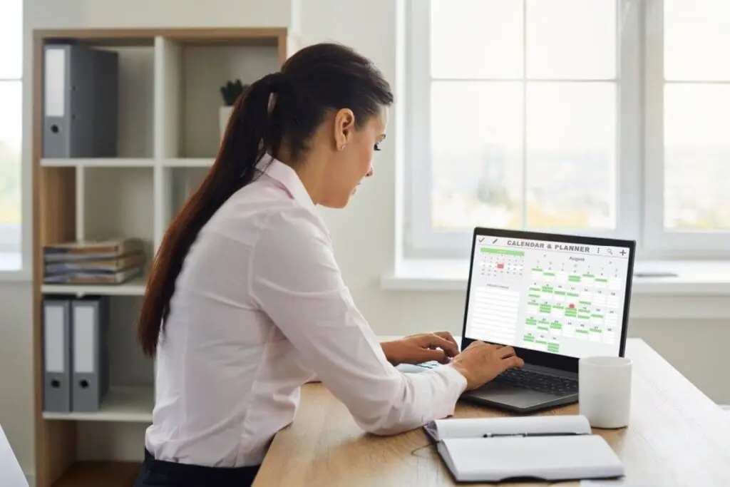 Mulher de camisa branca usando notebook com planner e calendário na tela, sentada em mesa de escritório iluminado por janelas