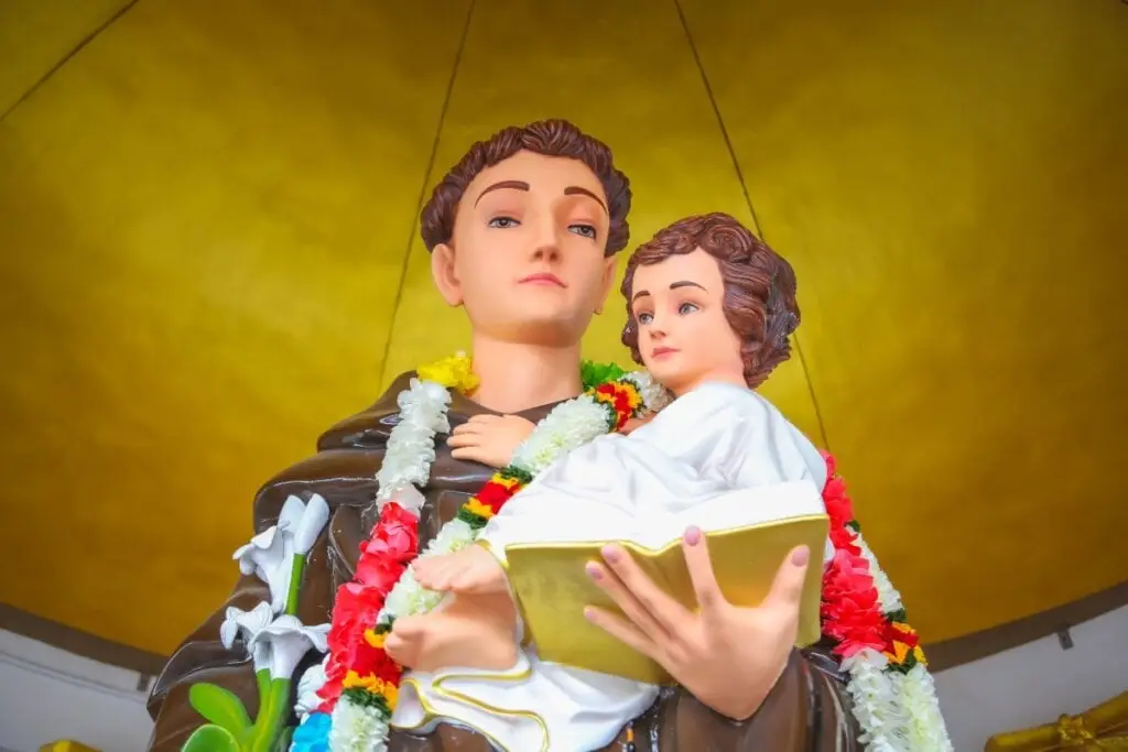 Uma estátua de Santo Antônio segurando o Menino Jesus, adornada com colares de flores coloridas, sob um fundo dourado