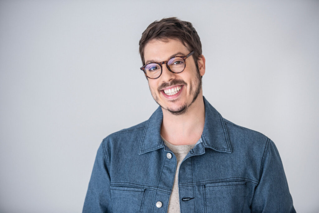 Homem com cabelo curto e barba, usando óculos preto e camisa jeans em fundo claro