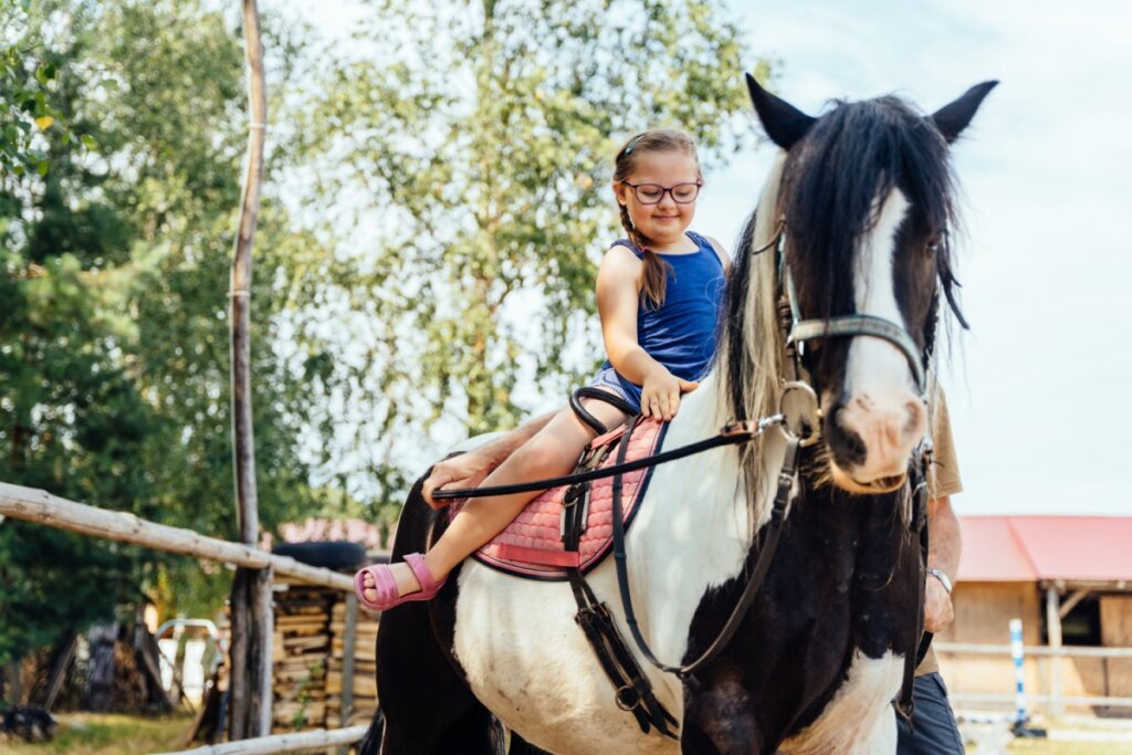 Menina com o cabelo preso em trança, usando regata azul, e short jeans e óculos andando a cavalo com ajuda de adulto 