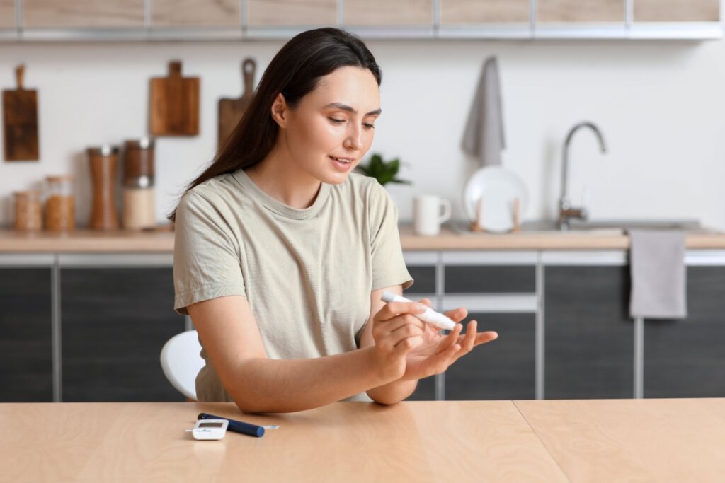 mulher de camiseta bege, sentada em mesa e espetando dedo com aparelho para medir glicemia