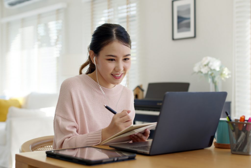 Menina de fone de ouvido estudando em frente a computador e realizando anotações, ela está usando um suéter rosa claro