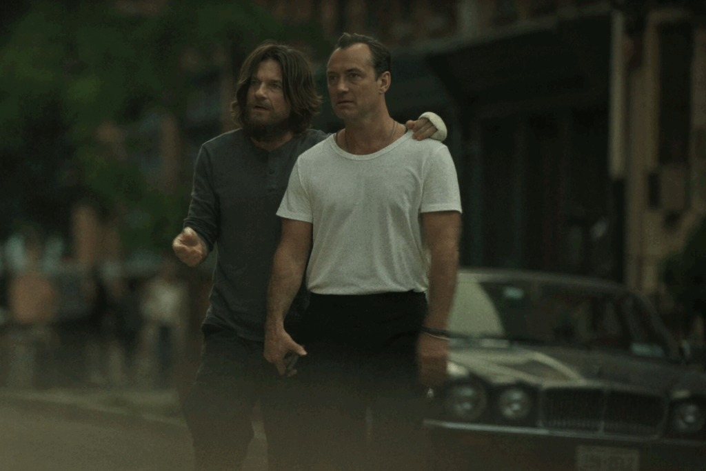 Dois homens juntos em uma rua, um deles com camiseta branca e o outro com barba e cabelo comprido usando camisa escura