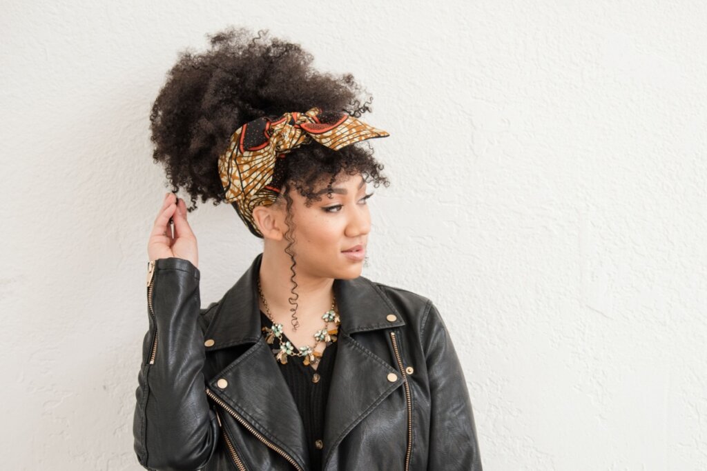 Jovem com o cabelo crespo preso em coque com lenço e usando jaqueta de couro e camiseta preta em fundo branco