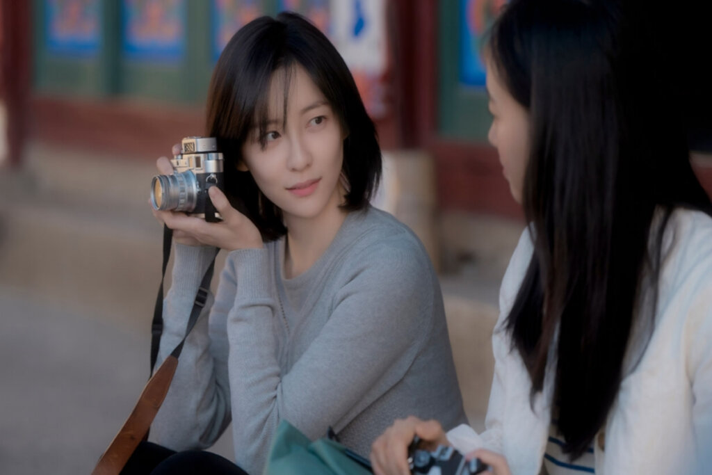 mulher de cabelo curto, blusa cinza e segurando câmera nas mãos. mulher de cabelo longo e camiseta branca ao lado dela