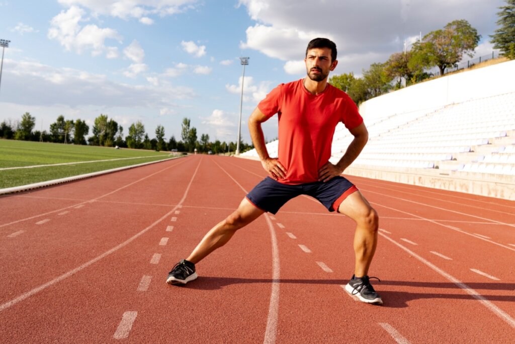 Homem de camiseta vermelha e shorts escuro alongando as pernas em pista de corrida com o chão vermelho