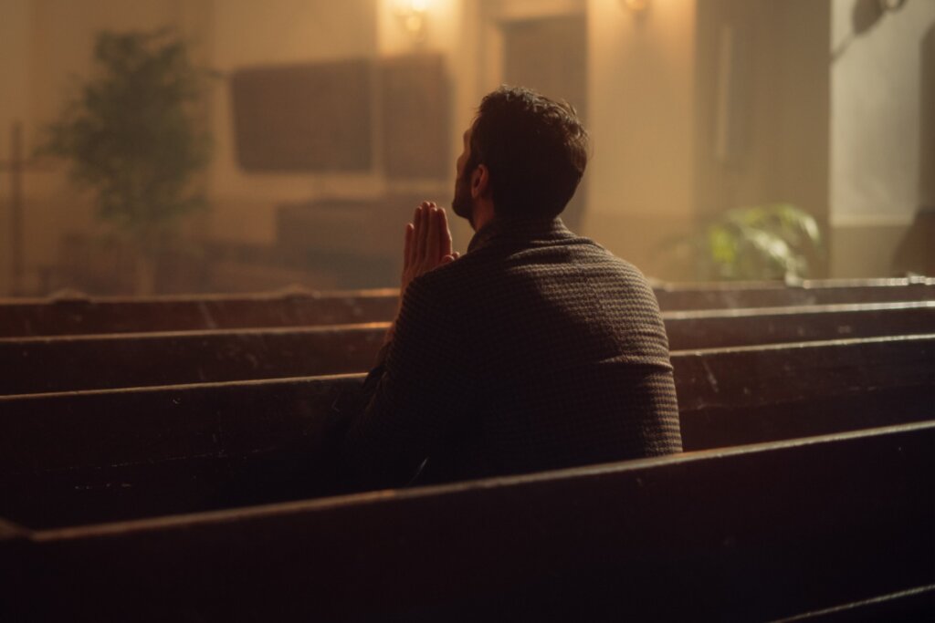 Homem usando camisa marrom orando sentado em banco de igreja
