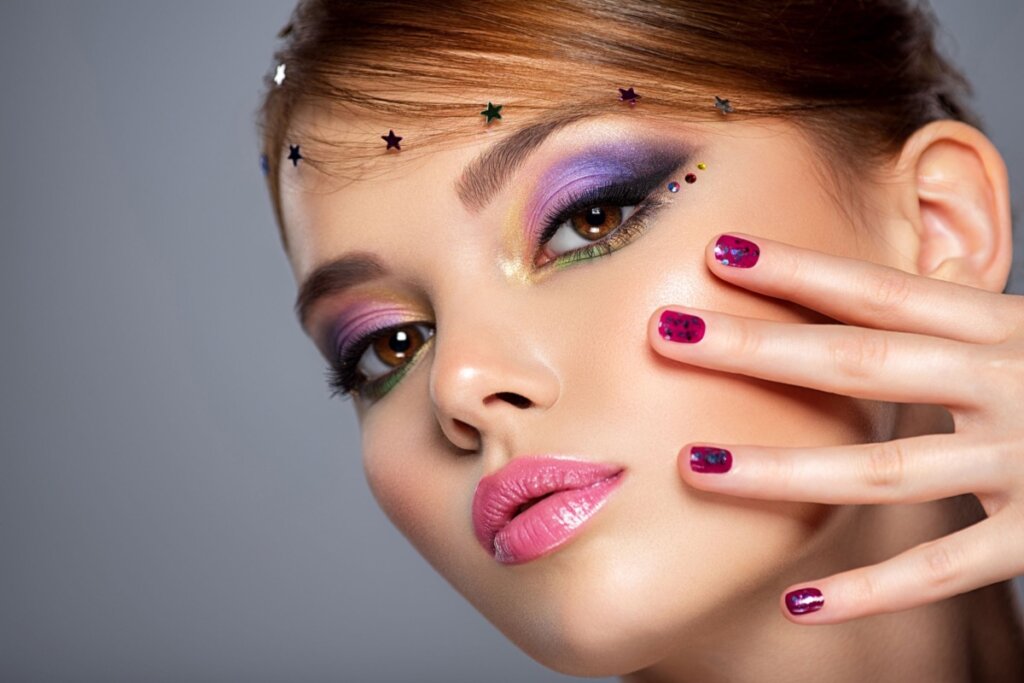Mulher com maquiagem colorida destacando sombras em roxo e rosa, batom rosa e unhas decoradas