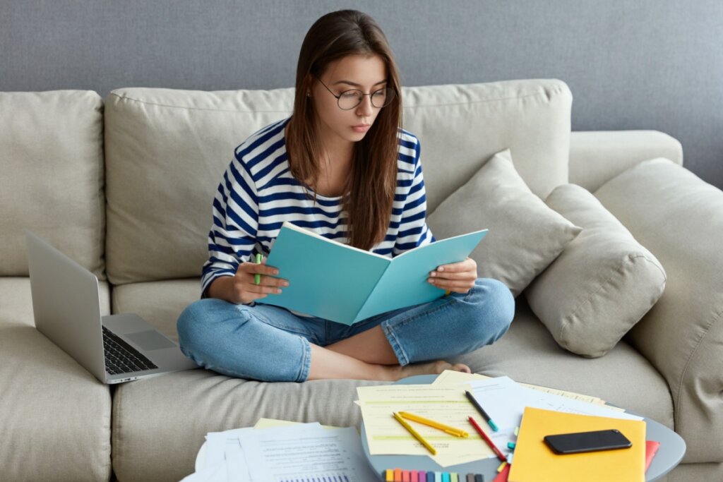 menina adolescente de blusa listrada e óculos, sentada em sofá com as pernas cruzadas e lendo livro com capa azul. mesa à frente dela com livros e canetas