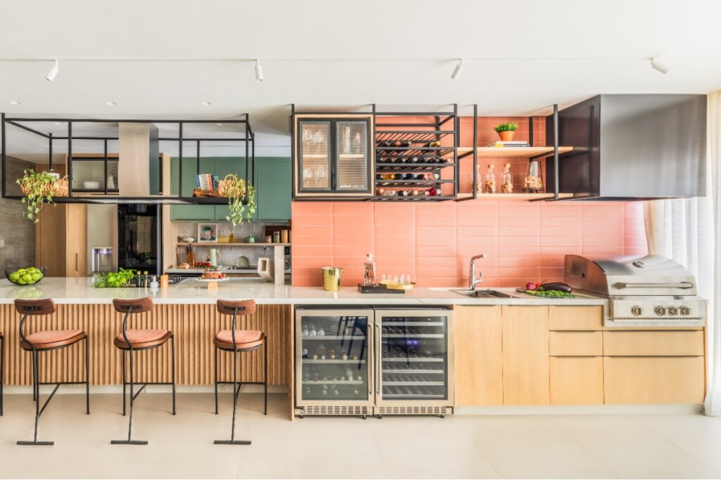 Cozinha gourmet com ilha ampla, banquetas altas, adega climatizada, churrasqueira embutida e prateleiras metálicas suspensas, com revestimento laranja no backsplash