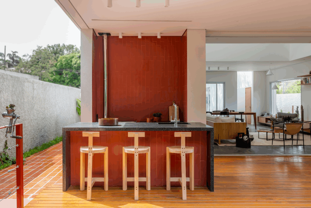Churrasqueira em área externa da casa com a parede revestida de piso vermelho e com banca em mármore preta com detalhes do mesmo revestimento vermelho da parede e três baquetas em madeira
