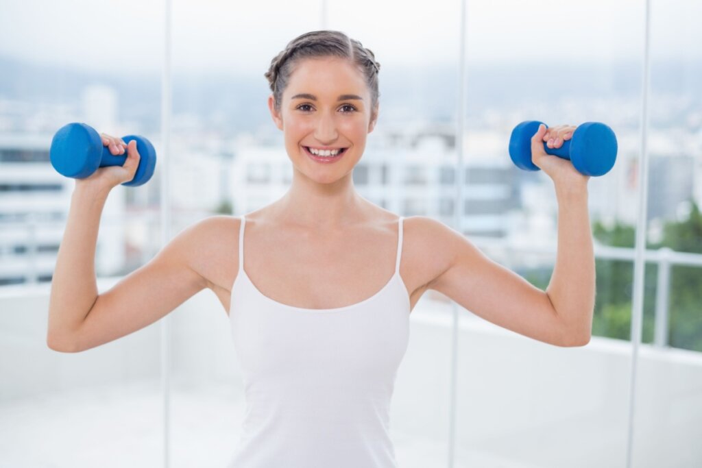 Mulher sorrindo e segurando dois pesos de academia. Cabelo com trança embutida e coque 
