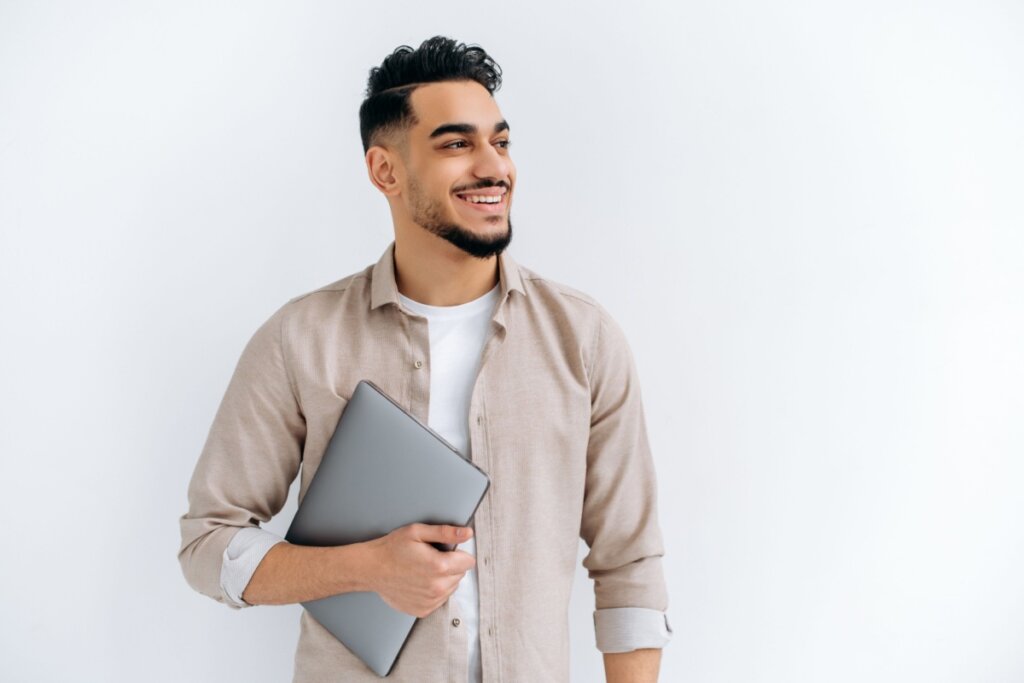 Homem com cabelo curto e barba feita em degradê usando camiseta branca e camisa de botões bege com um computador na mão em fundo claro