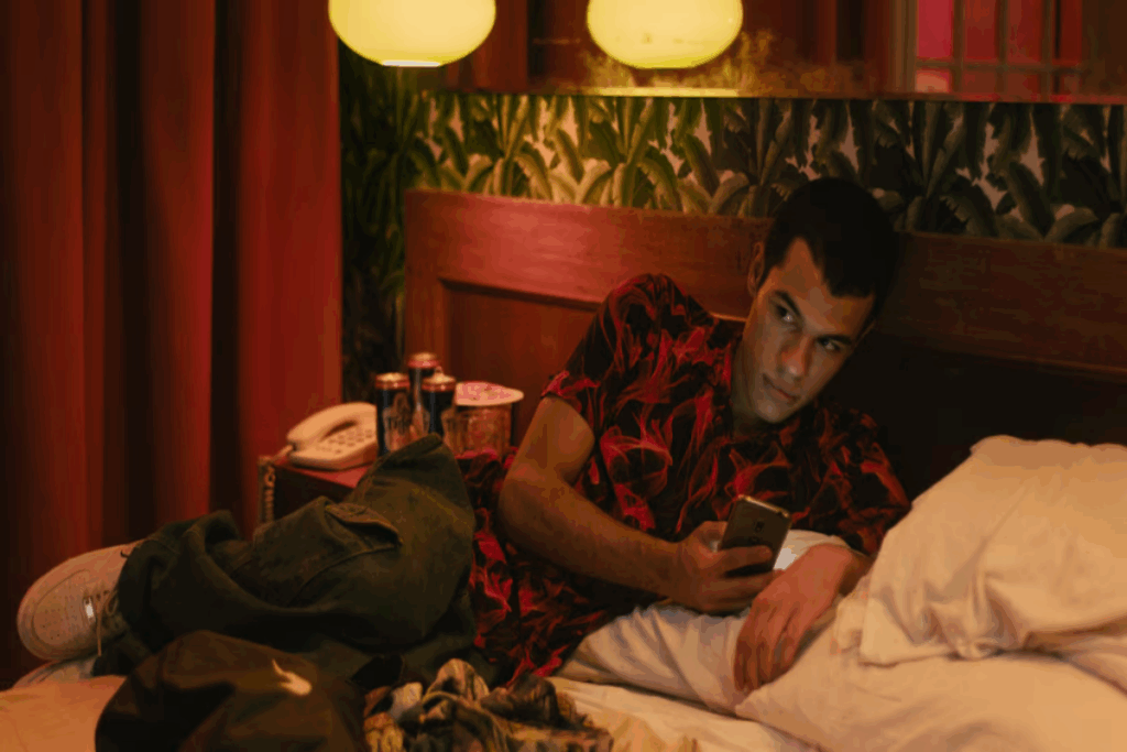 Homem com cabelo curto, usando camiseta estampada, calça jeans e tênis branco deitado na cama mexendo no celular em quarto de hotel 