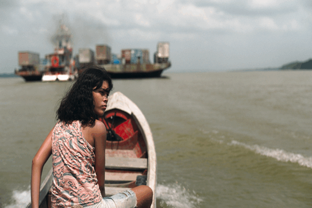 Uma jovem com cabelo curto, usando uma regata estampada e bermuda jeans andando em barco com barcos ao fundo