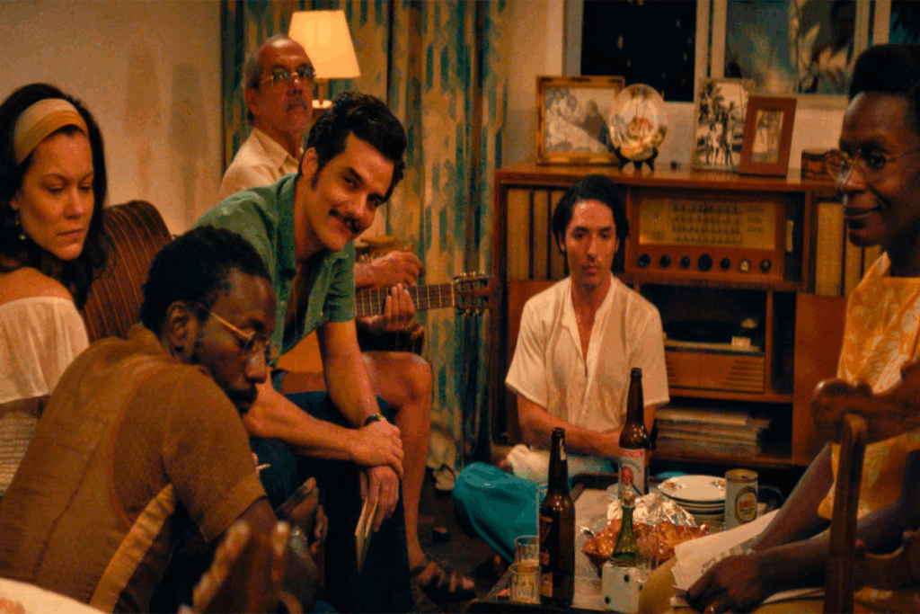 Grupo de pessoas sentada em sala ao redor de mesa com comida e bebidas, usando roupas da década de 70