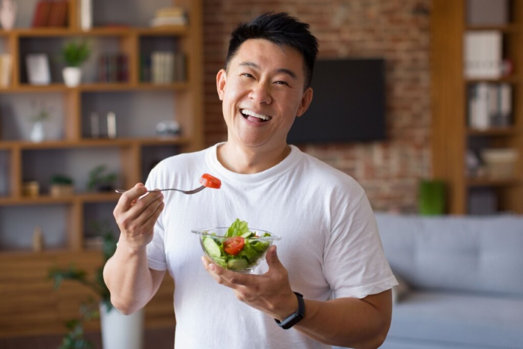 homem de camiseta branca sorrindo segurando garfo e pote de salada verde com tomate
