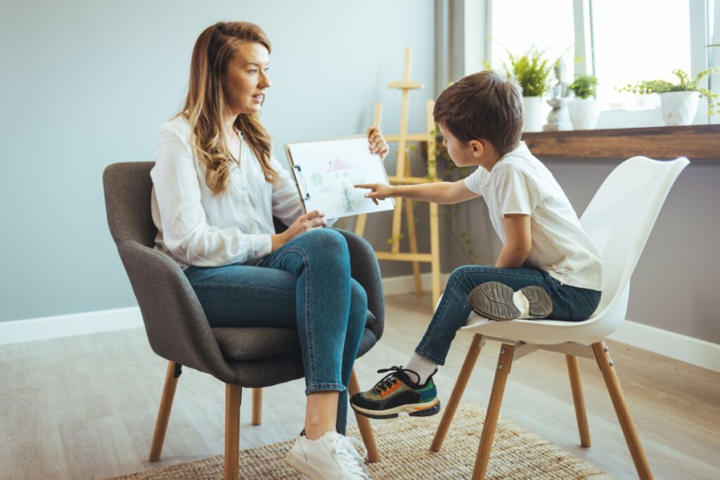 psicóloga e menino pequeno sentados em cadeira. ele aponta para papel na mão dela