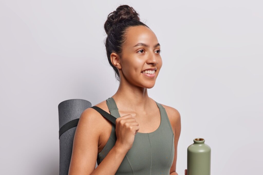 Mulher com penteado sleek bun olhando para o lado e segurando um colchonete e garrafa de água 