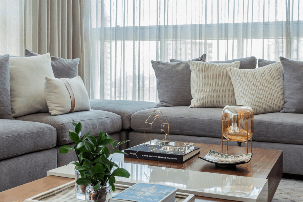 Sofá cinza com diversas almofadas cinza e bege com mesa de centro com itens de decoração e cortina branca ao fundo