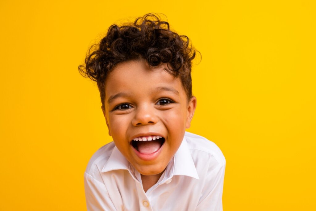 Menino com cabelo cacheado curto, usando camisa branca em fundo amarelo e sorrindo