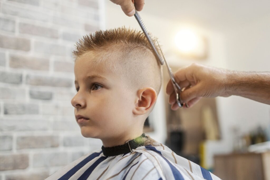 Mãos masculinas cortando o cabelo de menino no estilo moicano baixo em barbearia 