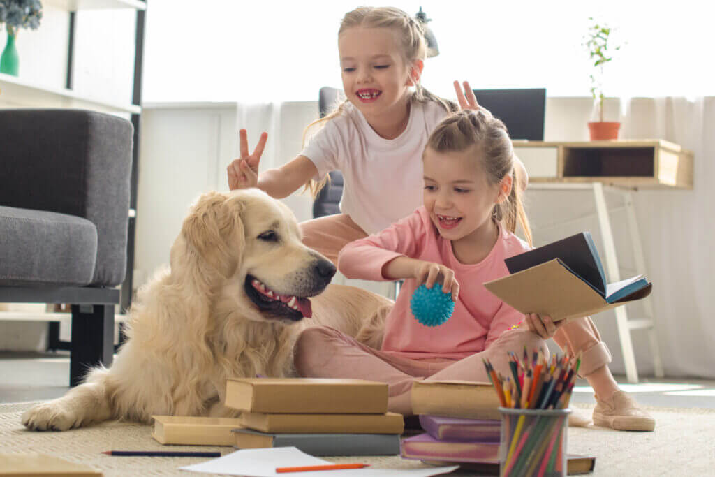 duas meninas sentadas no chão brincando com cachorro golden retriever bege com livros, bola azul e lápis