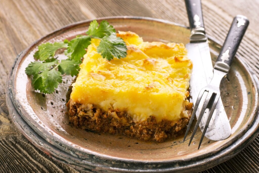 Um pedaço de escondidinho de carne moída em um prato de cerâmica bege claro, ao lado estão talheres e uma folha de cheiro verde