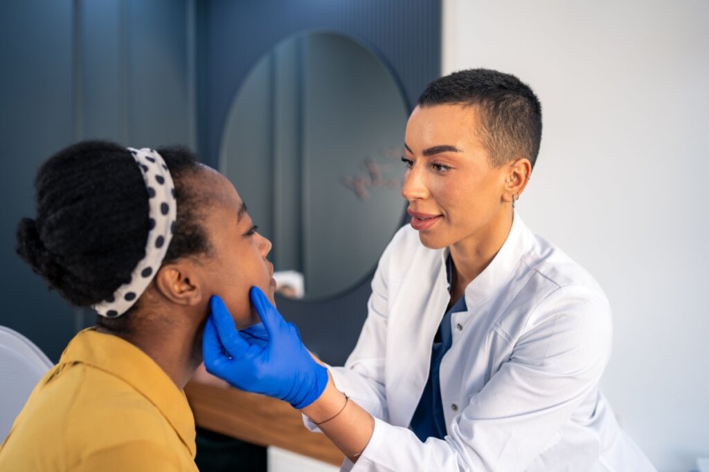 Jovem com o cabelo preso, usando faixa de bolinhas e camisa amarela sendo avaliada por dermatologista com o cabelo raspado usando jaleco branco e luvas azuis