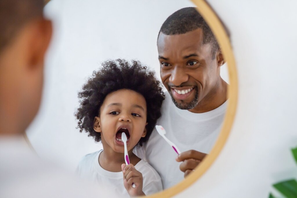 Pai com o cabelo curto e barba, orientando filho com o cabelo cacheado e solto a escovar os dentes em frente ao espelho, ambos estão usando camiseta branca