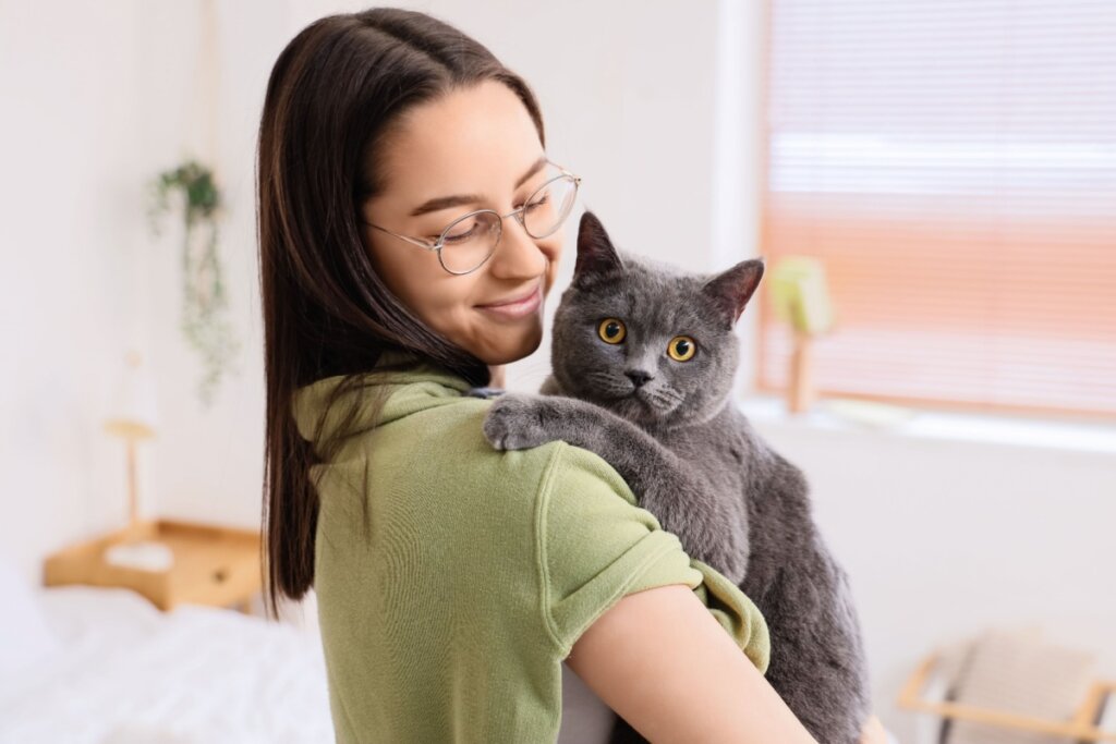 Mulher com cabelo liso e solto até o ombro, usando camiseta polo verde com gato com a pelagem cinza no colo
