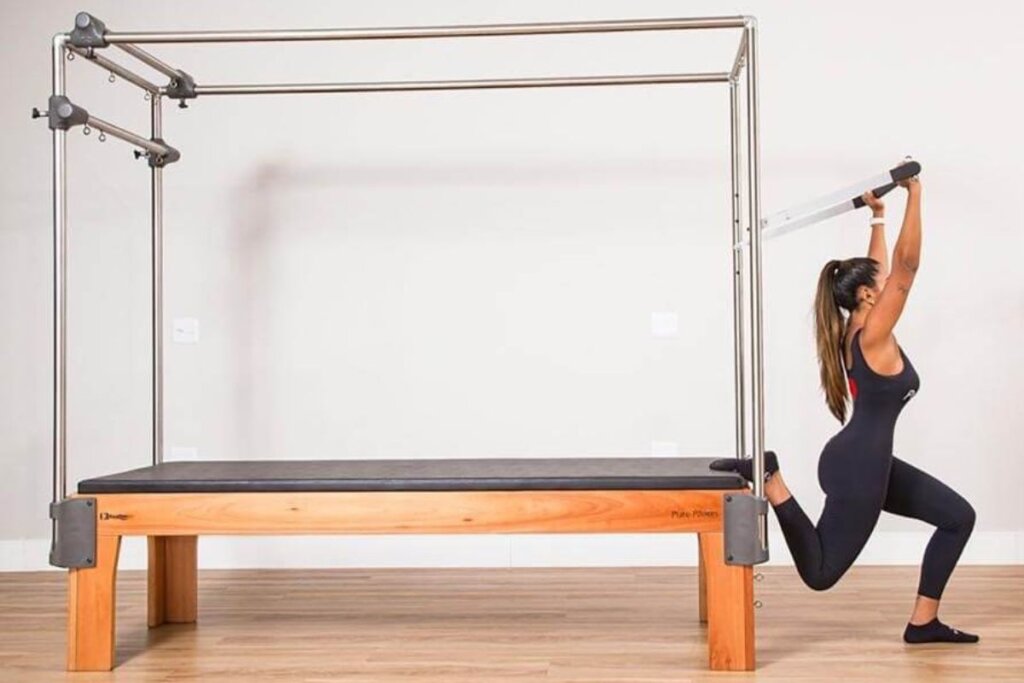 Mulher usando roupa preta, com uma das pernas apoiada em um cadillac de pilates, fazendo agachamento afundo