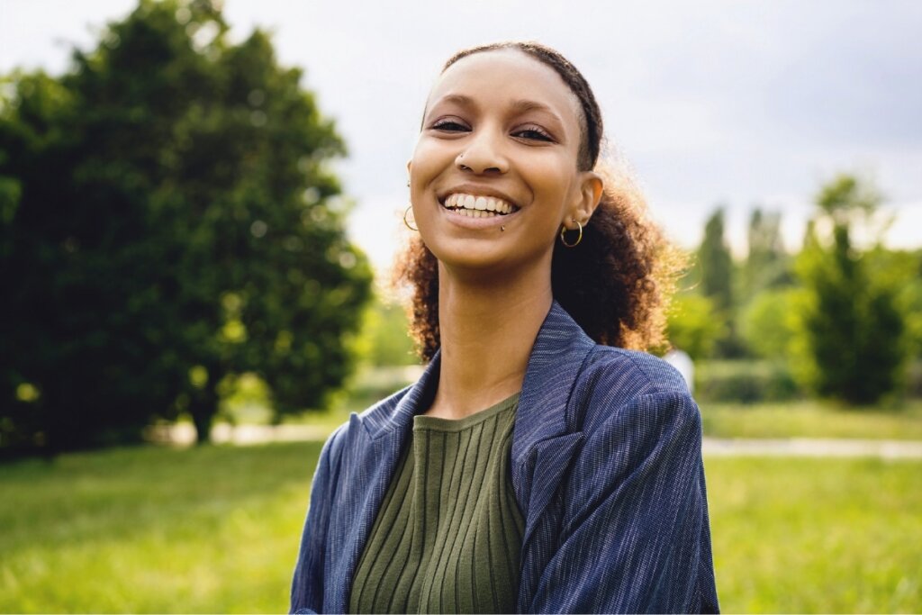 Mulher com cabelo cacheado com rabo de cavalo baixo, sorrindo em frente a um jardim