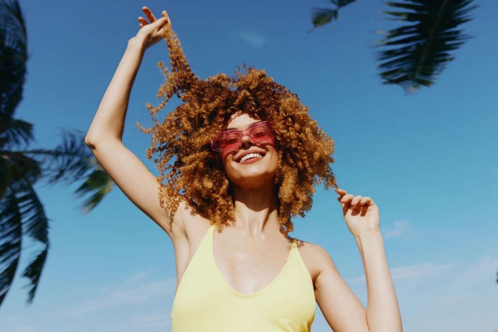 Mulher sorrindo, usando um óculos de sol e segurando mechas de cabelos em ambas as mãos