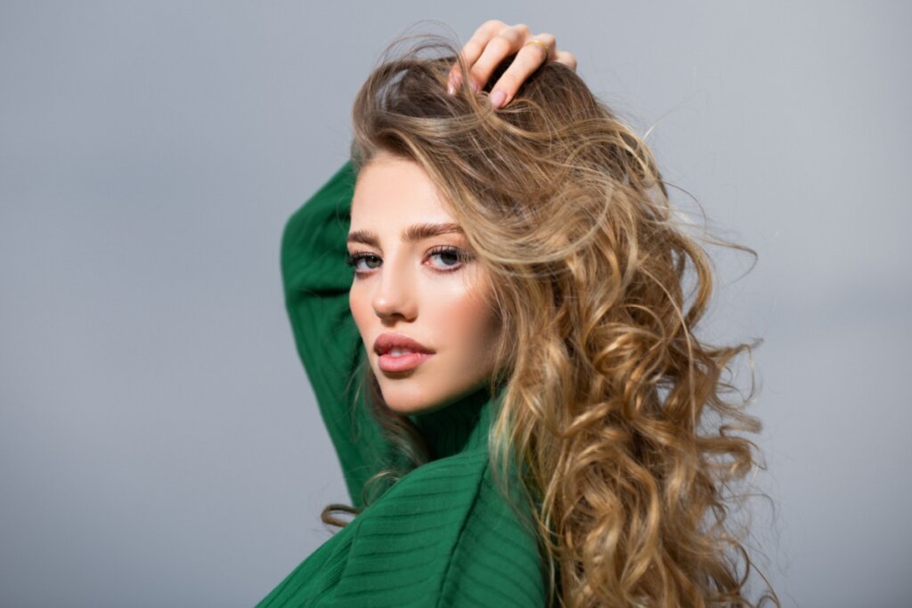 Mulher com o cabelo loiro mel usando suéter verde em fundo cinza e com a mão no cabelo