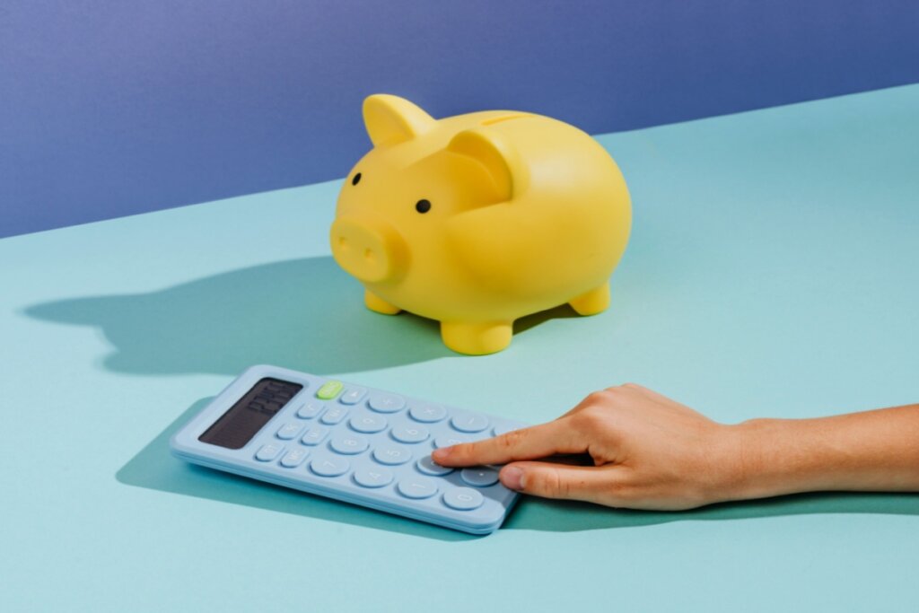 mão sobre calculadora azul ao lado de cofrinho amarelo em fundo azul-claro e escuro