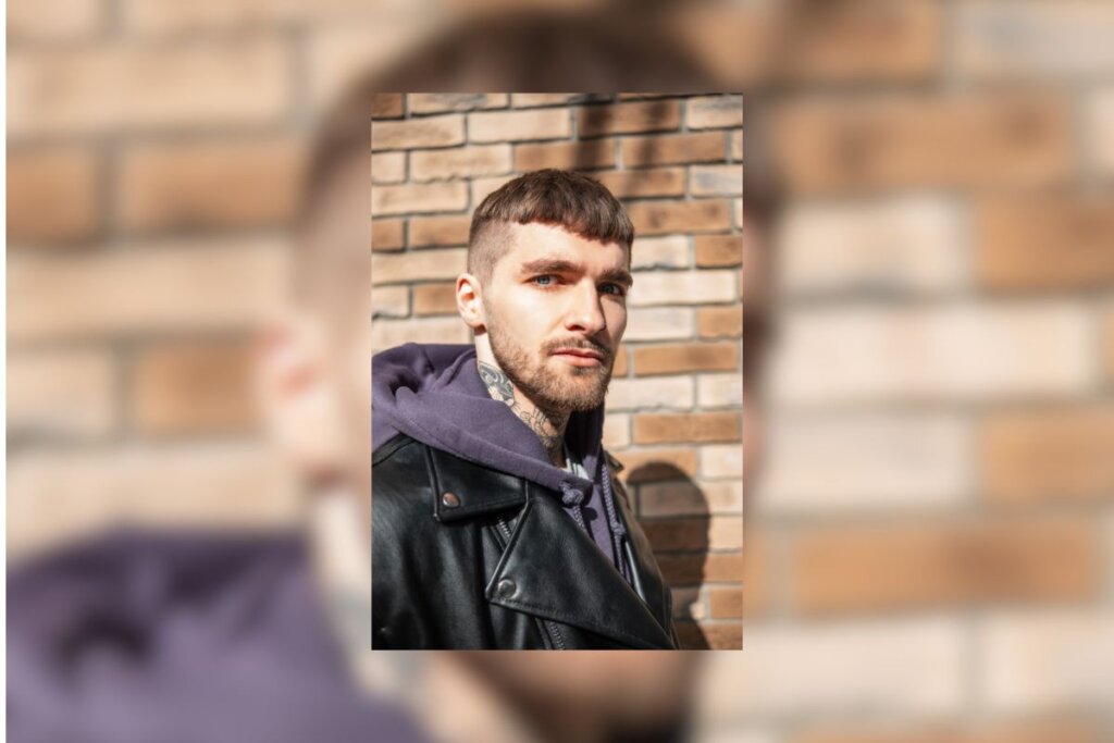 Homem com corte de cabelo caesar, usando uma jaqueta e em frente a uma parede de blocos