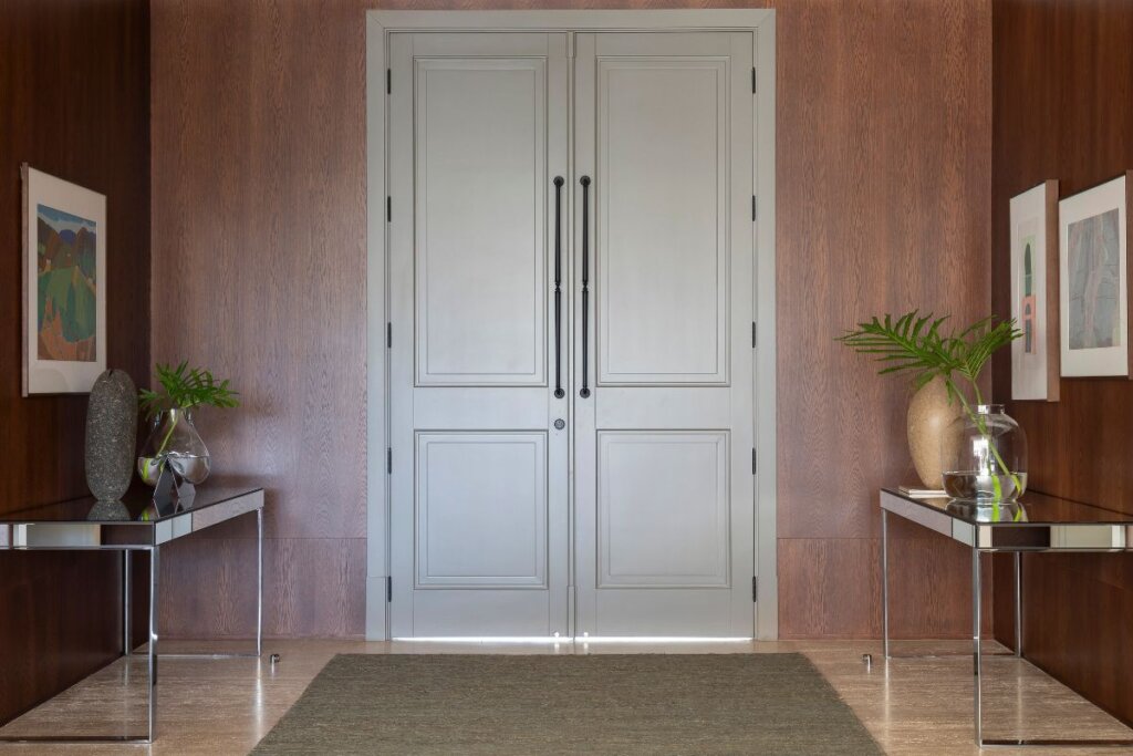 Hall de entrada com parede revestida de madeira e porta cinza claro. Ao lado, parede marrom com quadros e mesas de vidro em baixo