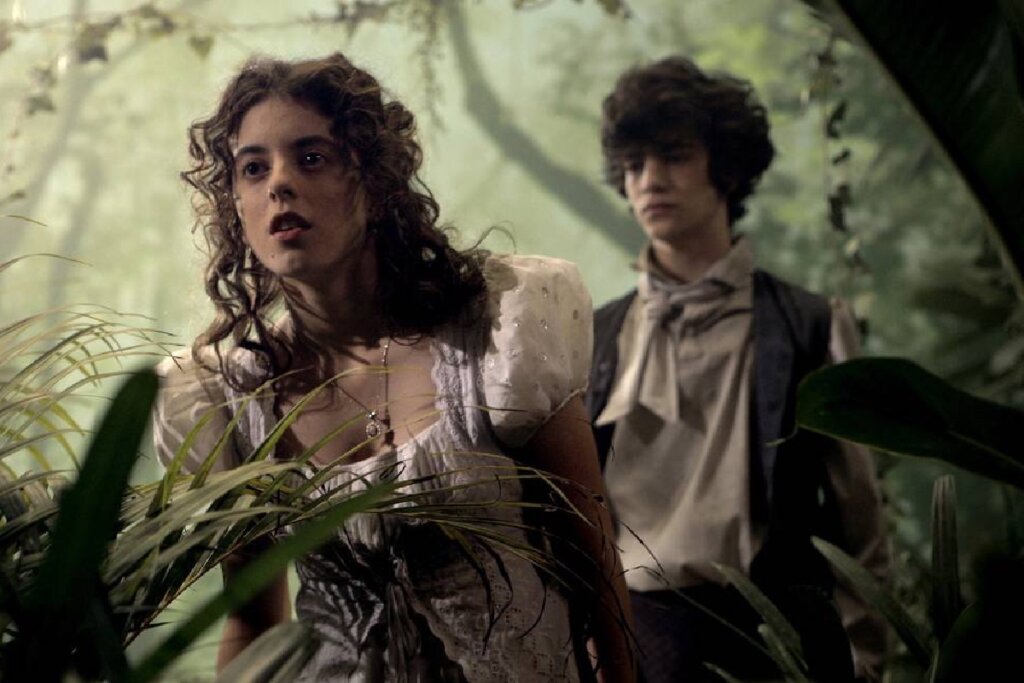 Dois jovens em roupas de época em meio à vegetação, com a garota em primeiro plano e o garoto ao fundo